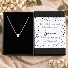 A personalized initial necklace with a silver pendant letter 'S' displayed in an open black gift box next to a matching card that reads, 'We couldn't have said 'I do' without you Sienna Thank you for being our flower girl.' The boxes are styled on a neutral fabric surface with dried flowers and green leaves.