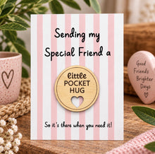 A pink and white striped greeting card with the text 'Sending my Special Friend a little POCKET HUG So it's there when you need it!' With removable wooden pocket hug token. displayed on a wooden surface with decorative plants and a small heart-shaped token.