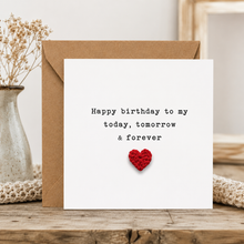 A square white birthday card with black typewriter-style text reading 'Happy birthday to my today, tomorrow & forever' and a small red crocheted heart near the bottom, shown with a brown kraft envelope on a wooden surface. Dried flowers in a vase and a knitted textile are visible in the background.