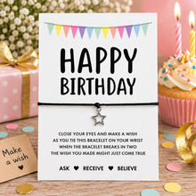 Happy birthday wish string bracelet. A white 'HAPPY BIRTHDAY' card with a black cord bracelet and silver star charm displayed on a wooden table, surrounded by birthday props including a cupcake, gift box, and confetti.