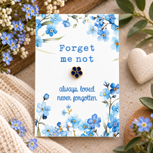 A floral memorial card with blue forget-me-not flowers and the words 'Forget me not' and 'always loved never forgotten,' featuring a small blue-and-gold flower-shaped pin centered on the card. The card is styled on a beige surface with knit fabric, greenery, and a small heart-shaped object around it.
