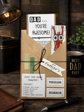 A Father's Day book gift set in a kraft paper envelope on a dark wooden table, featuring a 'DAD YOU'RE AWESOME!' card, a colorful monster card, a 'Just One more Chapter.' book, an index page marker, and a handwritten 'Father's' tag, with a black pen in the foreground.
