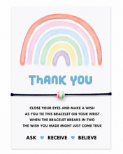 A square white card with a pastel rainbow illustration at the top, the words 'THANK YOU' in blue, and a thin black bracelet with a small striped rainbow bead across the centre. Smaller text below explains wish bracelet-wearing instructions with the words 'ASK RECEIVE BELIEVE' at the bottom.