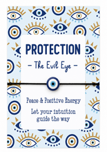 Holiday bracelet. A square card or box design featuring the words “PROTECTION - The Evil Eye -” on a white panel, surrounded by a light blue background patterned with navy and gold evil eye motifs. A dark cord bracelet with a matching evil eye bead runs horizontally across the centre, and additional text reads “Peace & Positive Energy” and “Let your intuition guide the way.