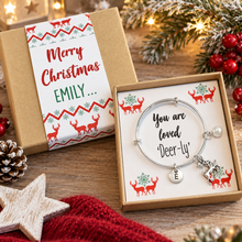 A personalised Christmas-themed gift set featuring a silver bangle bracelet with a 'You are loved ‘Deer-ly’' message and a silver letter 'E' charm, presented in an open kraft gift box. The matching gift box lid is shown closed with a white sleeve wrap that reads 'Merry Christmas EMILY. Christmas eve gift. Stocking filler.
