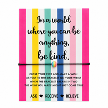 In a world where you can be anything. Be kind gift. A colourful card with a black cord wish bracelet across the centre. The card has vertical stripes in pink, white, yellow, teal, blue, and light pink, with a black inspirational quote and a small rainbow-striped charm bracelet. 