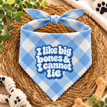 A blue and white gingham pet bandana with the text 'I like big bones & I cannot Lie' in white bubble letters with a blue outline, styled on a woven rope mat with dog toys and treats.
