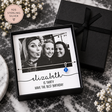 A personalised sterling silver necklace with a blue birthstone pendant is displayed in a black gift box with a black ribbon. The card inside features a black-and-white photo of three women and the text 'elizabeth IS THIRTY HAVE THE BEST BIRTHDAY'.