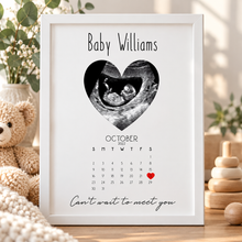 A personalised baby keepsake print in a white frame, featuring a black-and-white ultrasound heart graphic, the name 'Baby Williams,' an October 2022 calendar with a red heart on the 21st, and the phrase 'Can't wait to meet you.' The print is styled on a wooden surface with a teddy bear, plants, and wooden toys in the background.