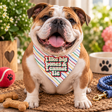 A smiling bulldog sits on a woven mat wearing a colorful striped bandana with the text 'I like big bones & I cannot lie'. The scene includes dog toys, bone-shaped treats, a bowl, and a leash in a bright, softly blurred outdoor setting.