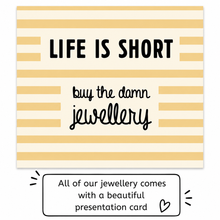 A square presentation card with cream and pale yellow horizontal stripes, featuring the text 'LIFE IS SHORT' in bold black letters and 'buy the damn jewellery' in black script. A lower callout box says, 'All of our jewellery comes with a beautiful presentation card' with a small heart icon.