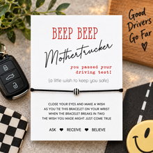 A white card with red and black text reading 'BEEP BEEP Mothertrucker you passed your driving test!' above a black cord bracelet with a silver center bead, styled on a tabletop with a car key, wooden sign, checkered cloth, and smiley-face sticker.