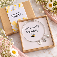 Personalised silver charm bangle with 3 charms. Don’t worry bee happy gift. A silver bracelet with bee, initial and pearl charms, presented in a kraft gift box with a white insert that reads 'Don't Worry Bee Happy.' The outside of the gift box is customised with the recipients name 
