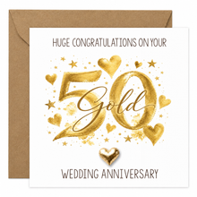 A square golden wedding anniversary card with a kraft brown envelope behind it. The card features watercolour gold text reading 'HUGE CONGRATULATIONS ON YOUR 50th WEDDING ANNIVERSARY' with gold hearts, stars, and a small gold heart keepsake at the bottom.