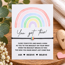 You got this wish string bracelet. A white card with a pastel rainbow illustration and the words 'You got this!' and a blessing, displayed on a wooden surface with flowers, greenery, and a small multicolor bracelet attached across the center.