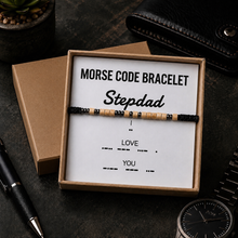A beaded Morse code bracelet presented in a brown gift box with a white card that reads 'MORSE CODE BRACELET Stepdad' and 'LOVE YOU' with dotted code. The box sits on a dark tabletop with a pen, wallet, and watch nearby.