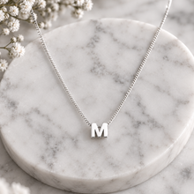 A silver initial necklace with the letter 'M' pendant, displayed on a round white marble tray with small white flowers in the corner.