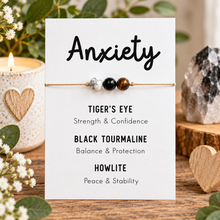 A white card displaying a beaded anxiety bracelet with tiger's eye, black tourmaline, and howlite beads, surrounded by decorative natural elements like a candle, flowers, and a crystal cluster.
