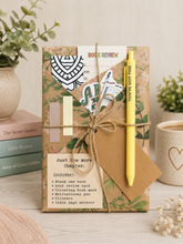 A wrapped book set tied with twine, featuring a yellow pen, stickers, and printed inserts, styled on a tabletop with books and small plants.