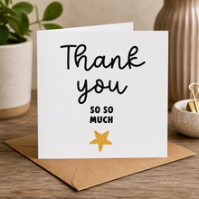 A white greeting card with the words 'Thank you SO SO MUCH' in black script and block lettering, plus a small yellow starfish illustration, displayed on a brown envelope on a wooden table with plants and home decor in the background.