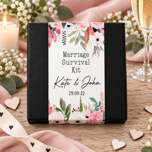 A black gift box with a floral wedding-themed label reading 'Marriage Survival Kit' and personalised names 'Kate & John' with the date '29.09. Customised wedding gift. Unusual wedding present. Personalised marriage survival kit. 1st anniversary present 