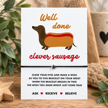 Clever sausage wish string bracelet. A white congratulations card with a dachshund in a hot dog illustration and the words 'Well done clever sausage,' tied with a black bracelet with a silver seat charm. Congratulations gift. Gift for new job or passing exams 