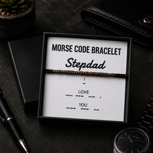 A beaded bracelet displayed inside a black gift box with a white insert that reads 'MORSE CODE BRACELET Stepdad' and additional Morse code words. The box is styled on a dark tabletop with a pen, wallet, and watch nearby.