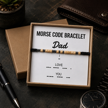 A Morse code bracelet gift set for Dad, featuring a beaded bracelet with black and natural wood beads displayed on a white card inside a brown box, surrounded by desk props including a wallet, pen, and watch.
