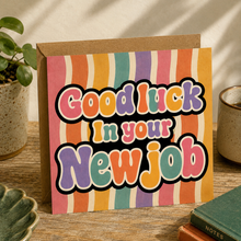 A square greeting card with a kraft envelope, featuring the text 'Good luck in your New job' in colorful retro bubble letters over vertical rainbow stripes. 