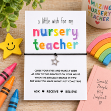 Nursery teacher wish string bracelet. A white card with colourful text reading 'a little wish for my nursery teacher' and a small silver star bracelet tied across the center, surrounded by nursery-themed props including crayons, a rainbow, a smiley face star, and a mug on a wooden surface.