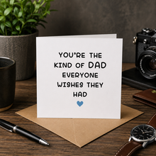 A white greeting card with black text reading 'YOU'RE THE KIND OF DAD EVERYONE WISHES THEY HAD' and a small blue heart, displayed on a kraft envelope on a wooden desk with a plant, camera, pen, watch, and wallet around it.