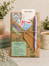 date night with a book tied with twine, displayed on a tabletop with books and a mug in the background. The kit includes a decorative card, stickers, a pen, and a green label reading 'Just One more Chapter.