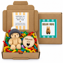 A teddy bear gift box set featuring a plush teddy bear wearing a gray knit hat, nestled in colorful shredded paper inside an open brown cardboard box with a wooden 'little pocket HUG' tag. A matching closed brown box with a 'BEAR HUG' label sits behind it on a white background.