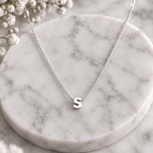 A silver chain necklace with a small lowercase letter 's' pendant, displayed on a round white marble tray with small white flowers in the corner.