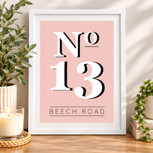 New home gift. A framed typographic art print leaning on a wooden surface, with a pink background, large white 'No 13' lettering, and 'BEECH ROAD' beneath it. The white frame is styled in a home setting with a candle, potted plant, and books.