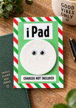 A novelty gift card featuring a white circular felt patch with googly eyes, bordered by red and green stripes, with the text 'iPad' and 'CHARGER NOT INCLUDED'. The card is styled on a wooden desk next to a green notebook, a pen, and a mug that reads 'GOOD VIBES ONLY'.