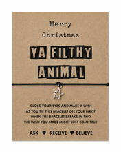 A Christmas-themed bracelet set featuring a brown kraft paper background with black text reading 'Merry Christmas YA FILTHY ANIMAL' and a black cord bracelet with a small silver star charm hanging across the middle. Stocking filler gift. Christmas bracelet.