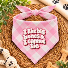 Pink and white gingham pet bandana with a bow, featuring the text 'I like big bones & I cannot lie' in raised bubble letters, styled on a woven placemat with dog treats and toys.