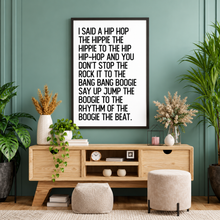 A framed typographic wall art print of Rapper’s Delights lyrics with black text on a white background hangs above a light wood media console in a styled room with green plants, poufs, and decor.