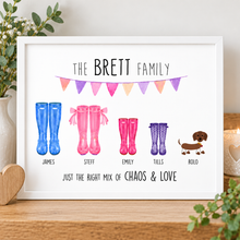 A framed personalised welly family print on a wooden surface, featuring the text 'THE BRETT FAMILY' and 'JUST THE RIGHT MIX OF CHAOS & LOVE' with colourful illustrated rain boots and a dachshund labeled 'ROLO'.