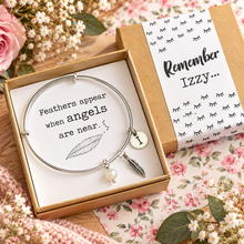 Personalised silver charm bangle bracelet with feather charms and a pearl bead is presented in a kraft gift box with a printed message that reads feathers appear when angels are near. Personalised on the outside with the recipients name.