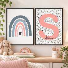 Girls room print set. Nursery wall art. Two framed personalised nursery art prints displayed side-by-side on a wooden shelf in a styled nursery setting. The left print features a rainbow and the right one letter S in peach pink with the name 'sienna' written vertically in black script on a white background with black polka dots.