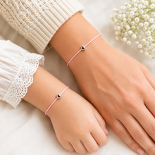 Matching mother daughter wish string bracelets. Two delicate pink cord bracelets with small silver heart charms worn on a person's wrists, styled with a cream knit sweater and white lace cuffs, with small white flowers in the background.