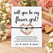 Will you be my flower girl proposal wish string bracelet. A white card with black script text reading 'will you be my flower girl?' and a black cotton heart charm bracelet across the centre, styled with a small floral wreath and heart charm.