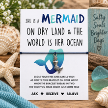 A mermaid-themed bracelet gift set. The card reads 'SHE IS A MERMAID ON DRY LAND & THE WORLD IS HER OCEAN' and shows a small silver mermaid charm tied on a black cotton string bracelet. Mermaid wish string bracelet. 