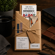 A kraft paper book package for daddy tied with twine sits on a wooden table surrounded by lifestyle props including a black mug, a potted plant, a leather journal, and glasses. The package contains a book or booklet with printed inserts reading 'Some People Don't Believe in Superhero's But They Haven't Met My Daddy' and 'Just One More Chapter: Includes:'.