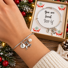 A personalised Christmas silver charm bracelet worn on a person's wrist, featuring a deer charm, a 'me' charm, and a pearl. The background shows a gift box with the message 'You are loved Deer-ly' and festive Christmas decorations. Secret santa gift.