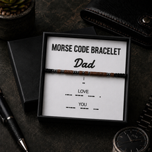 A beaded Morse code bracelet presented in a black gift box with a white card that reads 'MORSE CODE BRACELET Dad' and additional coded words below, styled on a dark tabletop with a pen, wallet, plant, and watch nearby.