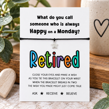 A black cord bracelet with a small silver star charm displayed on a white card with a humorous retirement message. The card is styled on a wooden surface with green leaves and a ceramic mug in the background.