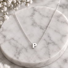 A silver initial necklace with a lowercase-style letter 'p' pendant, displayed on a round white marble tray with small white flowers in the corner.